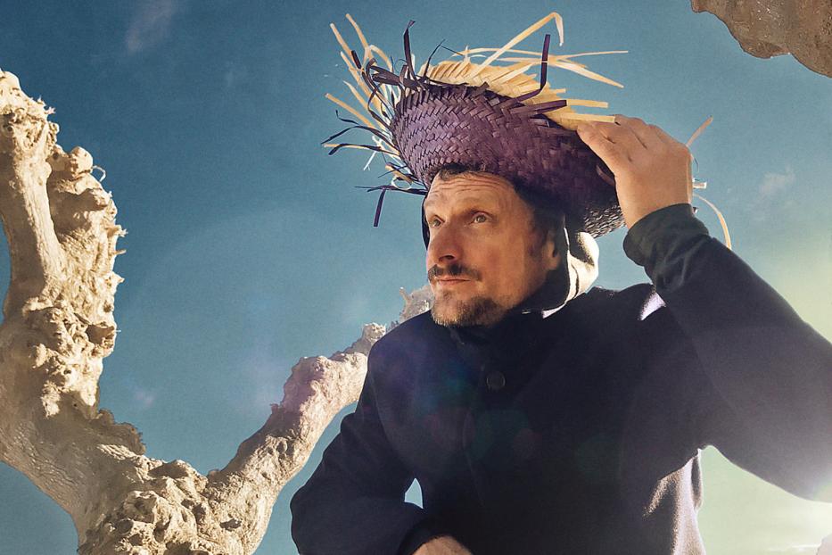 Ein Mann mit Bart trägt einen ungewöhnlichen Hut vor blauem Himmel.