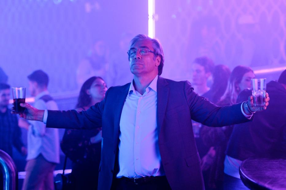 Javier Bardem steht mit ausgestreckten Armen und je einem Drink in der Hand in einem Club.