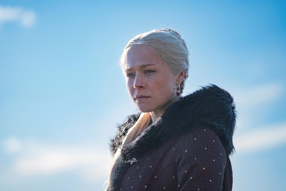 Rhaenyra Targaryen mit blonden Haaren und dunklem Umhang vor blauem Himmel.