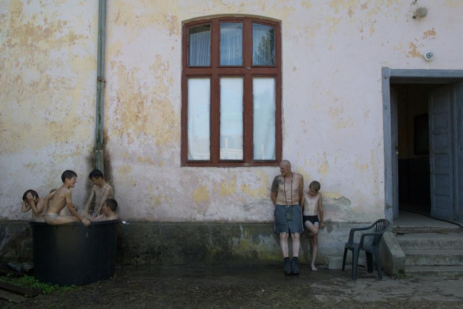 Eine Gruppe Kinder badet in einem Bottich vor einem Haus, während ein Mann zusieht.