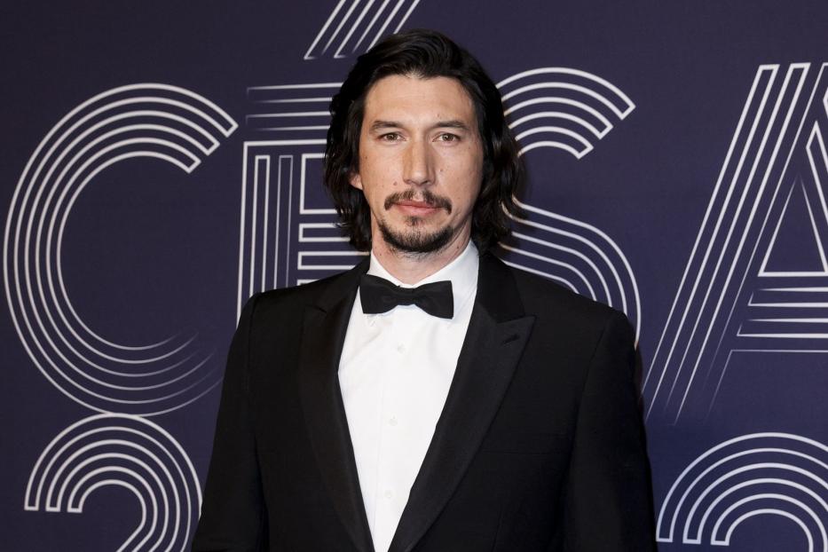 Adam Driver in einem schwarzen Smoking vor einem grafischen Hintergrund.