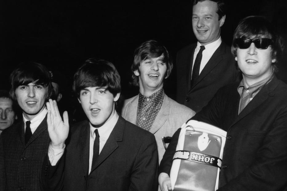 Die Beatles posieren mit einer Tasche mit der Aufschrift „John“ für ein Schwarzweißfoto.