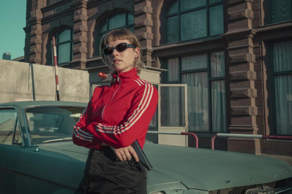 Eine Frau mit Sonnenbrille und roter Jacke lehnt an einem Oldtimer und hält eine Pistole.