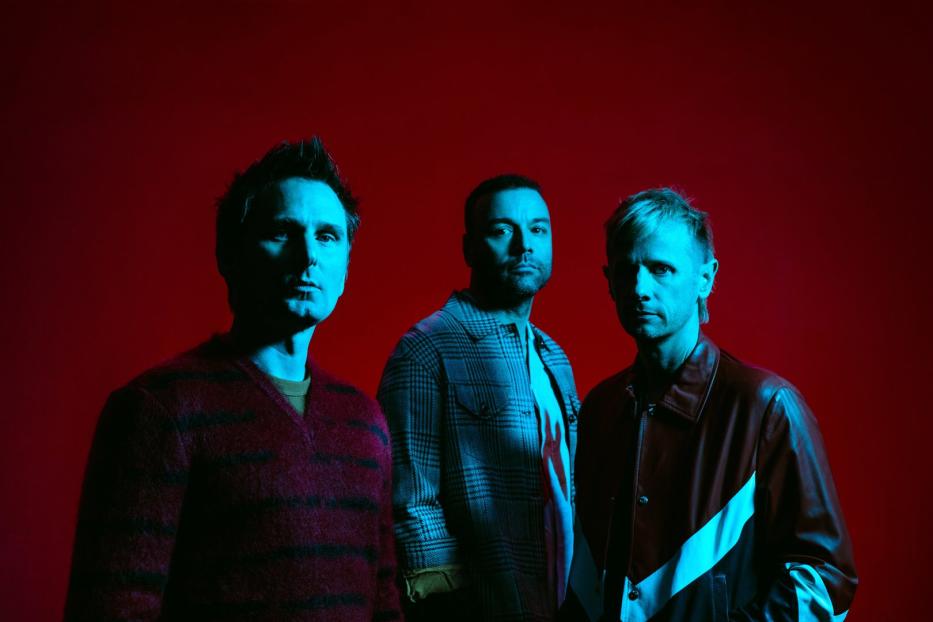 Die Band Muse posiert vor einem roten Hintergrund.