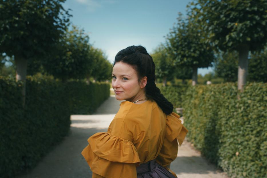 Eine junge Frau in historischer Kleidung blickt in einem Garten über ihre Schulter.
