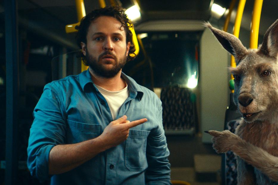 Ein Mann und ein Känguru sitzen in einem Bus und zeigen mit dem Finger in die gleiche Richtung.