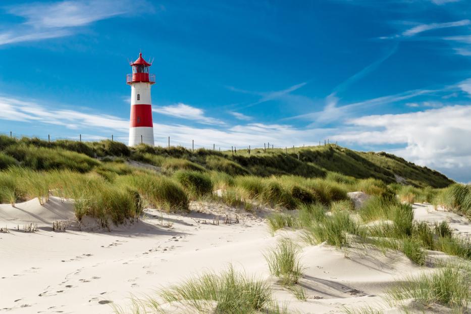 Ein rot-weiß gestreifter Leuchtturm auf einer Düne mit Strandhafer unter blauem Himmel.