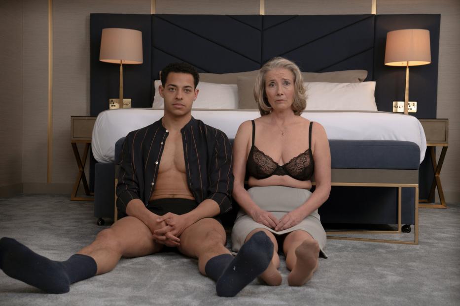 Ein junger Mann und eine ältere Frau sitzen in Unterwäsche vor einem Bett.