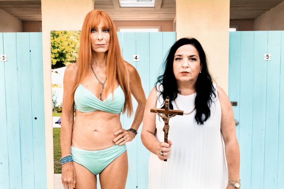 Zwei Frauen posieren vor hellblauen Strandkabinen, eine im Bikini, die andere mit einem Kruzifix.