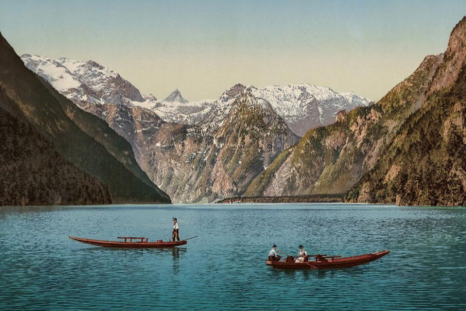 Zwei Boote fahren auf einem See vor einer Bergkulisse mit schneebedeckten Gipfeln.