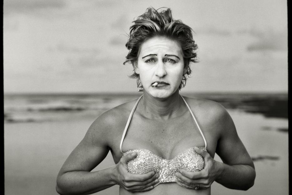 Eine Frau mit Clownsgesicht und glitzerndem Bikini-Oberteil am Strand raucht eine Zigarette.