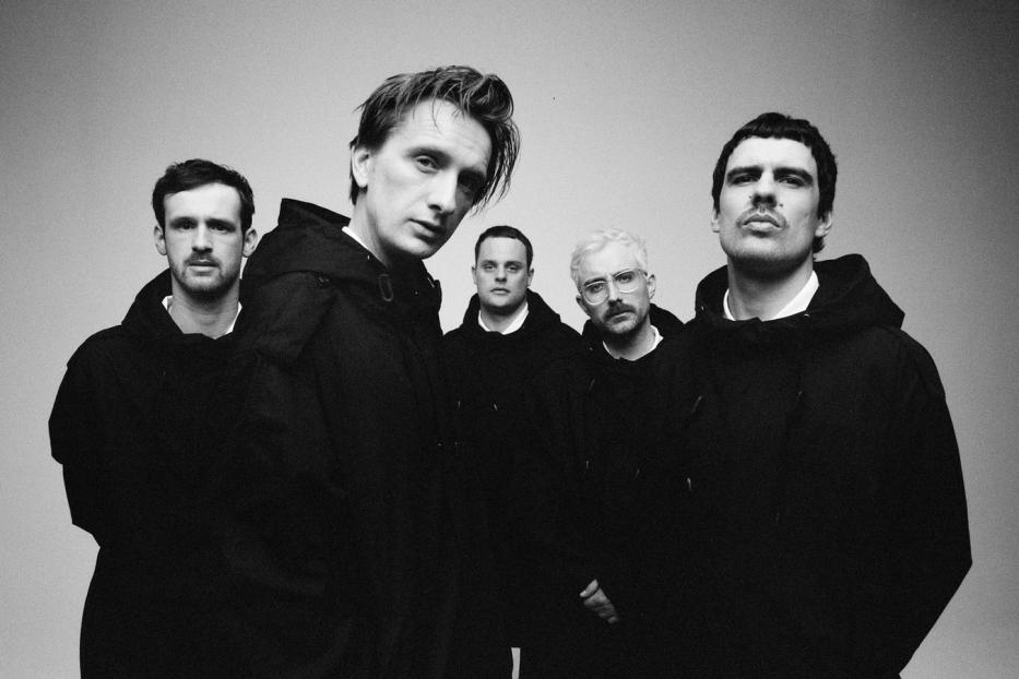 Schwarzweißaufnahme einer Band, die schwarze Kapuzenpullover vor einem neutralen Hintergrund trägt.