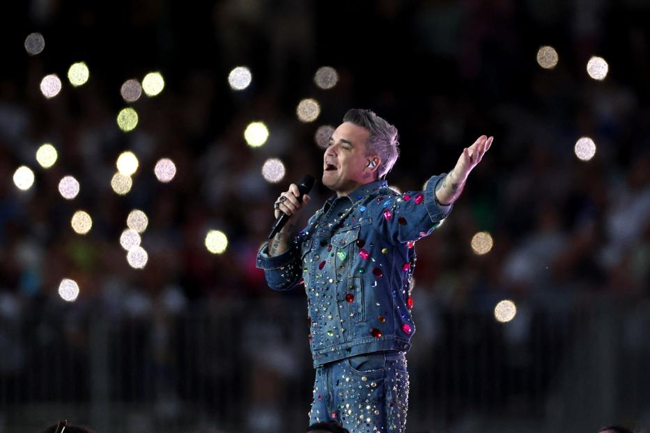 Robbie Williams tritt in einer mit bunten Juwelen besetzten Jeansjacke auf einer Bühne auf.