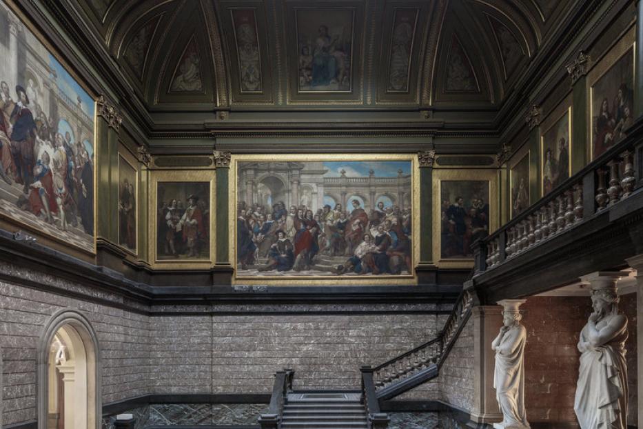 Ein weitläufiger Museumssaal mit Gemälden, Statuen und einer kunstvollen Treppe.