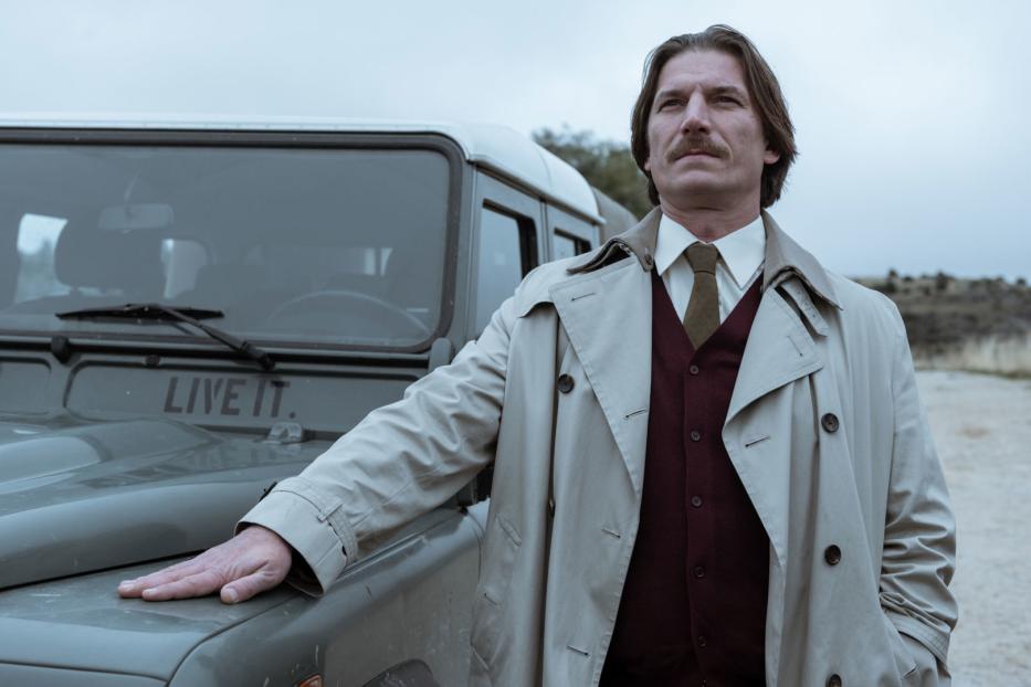 Ein Mann mit Trenchcoat und Schnurrbart lehnt an einem Geländewagen, auf dem „Live it.“ steht.