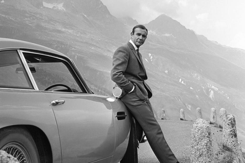 Sean Connery lehnt an einem Aston Martin vor einer Bergkulisse.