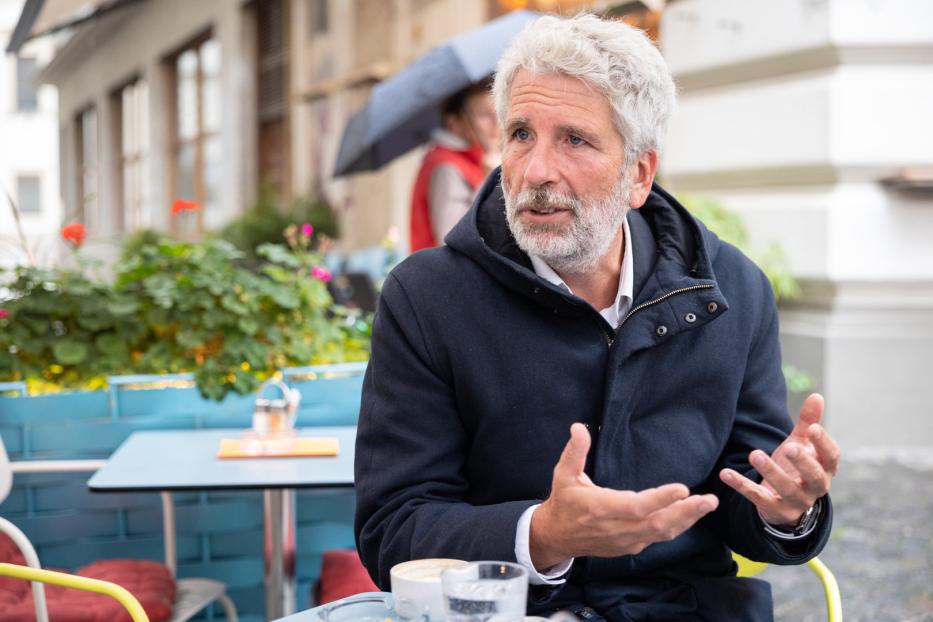Ein Mann mit grauem Haar gestikuliert an einem Tisch im Freien.