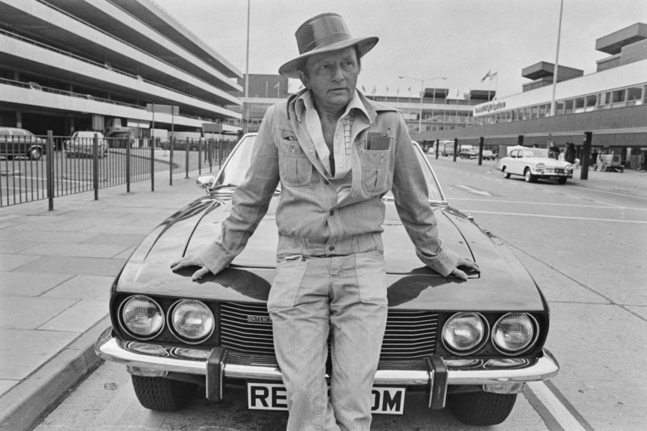 Ein Mann mit Hut lehnt an einem Jensen Interceptor vor einem Flughafengebäude.