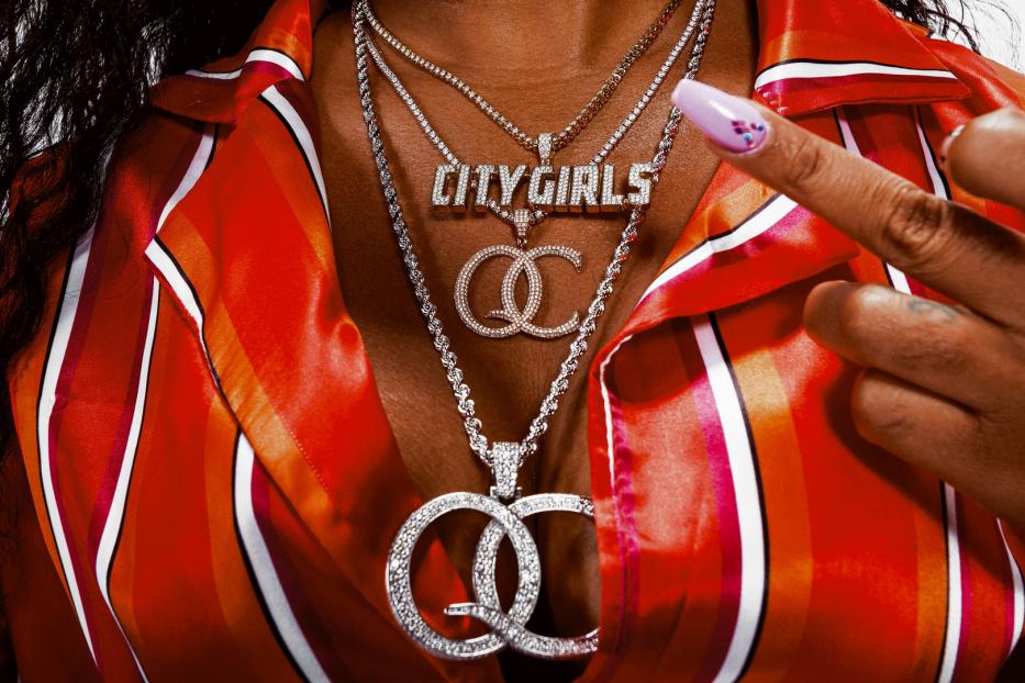 Nahaufnahme einer Person mit mehreren Halsketten, darunter eine mit der Aufschrift „City Girls“.