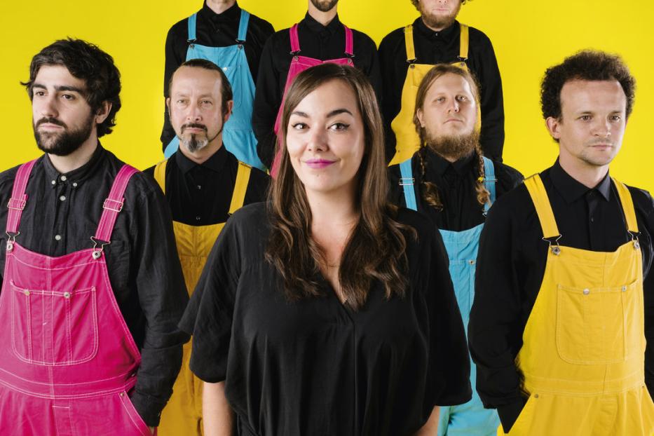 Eine Gruppe von acht Personen vor einem gelben Hintergrund, gekleidet in farbenfrohe Overalls.