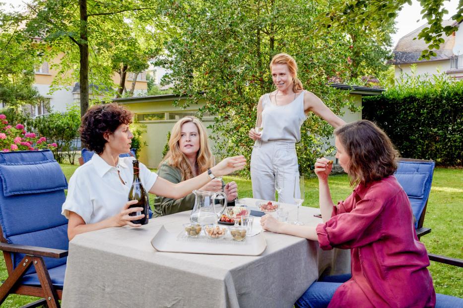 Vier Frauen sitzen im Garten an einem Tisch mit Getränken und Snacks.