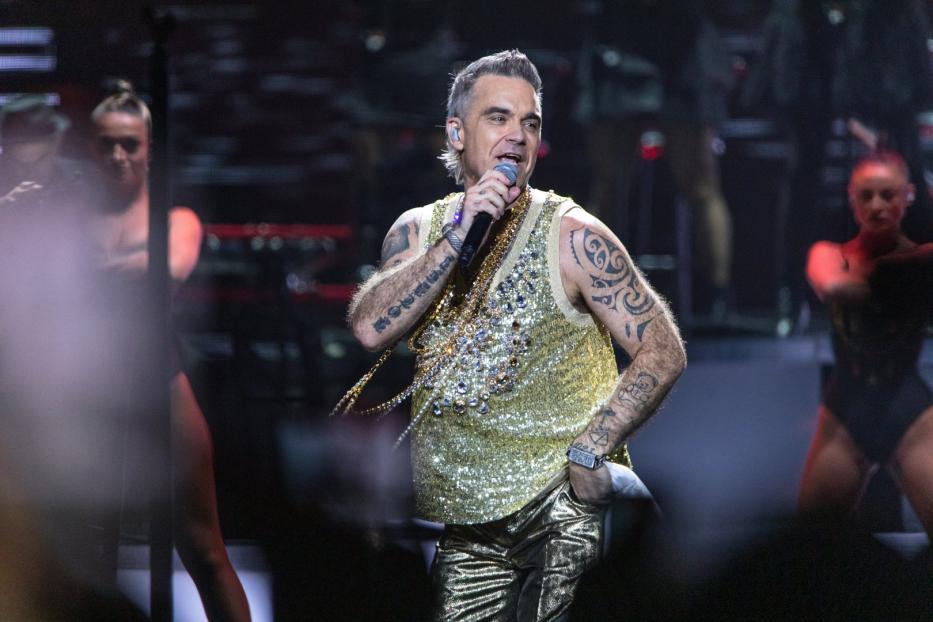 Robbie Williams tritt in einem goldenen Glitzertop und goldenen Hosen auf der Bühne auf.