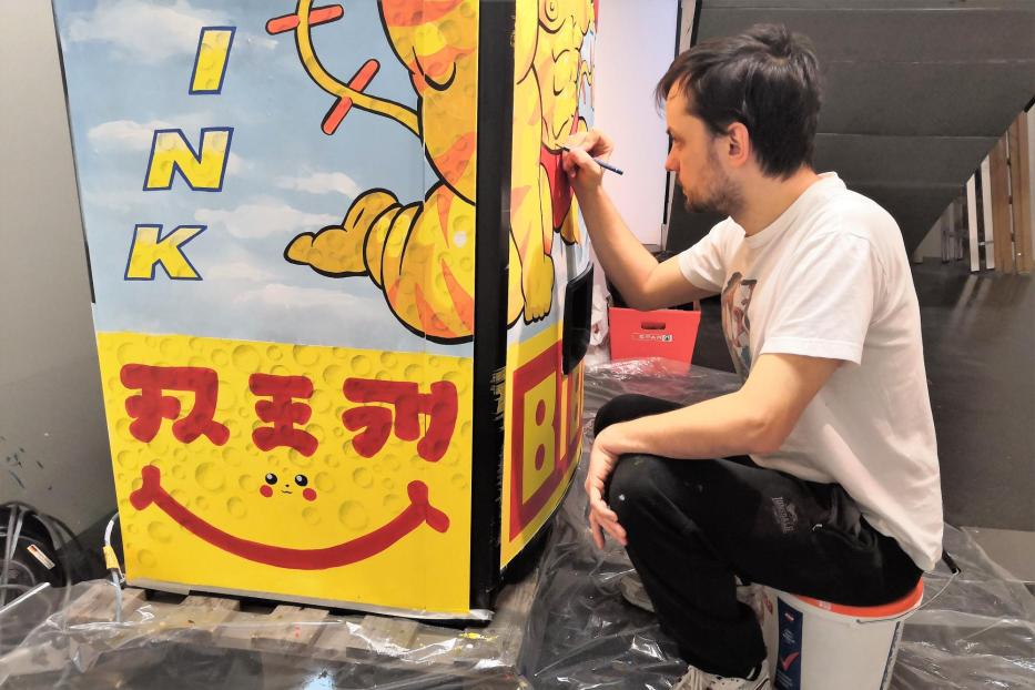 Ein Mann bemalt eine bunte Werbetafel mit der Aufschrift „INK“ und japanischen Schriftzeichen.