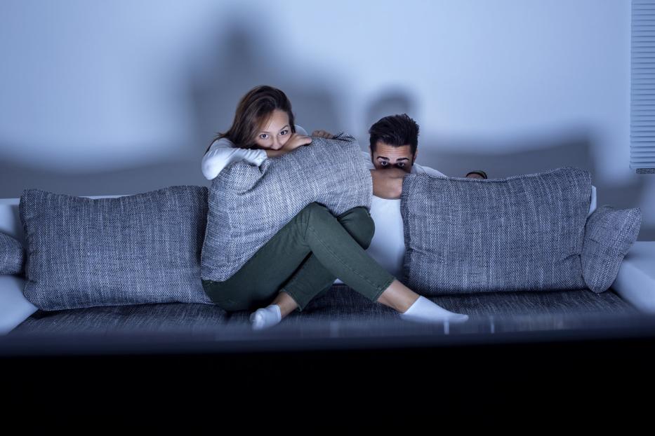 Ein Paar versteckt sich ängstlich hinter Kissen auf einem Sofa.