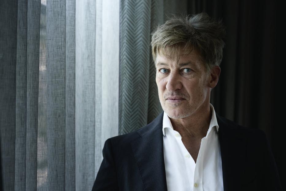 Ein Mann mit blau-grauen Augen und grau meliertem Haar trägt ein weißes Hemd und einen schwarzen Blazer.