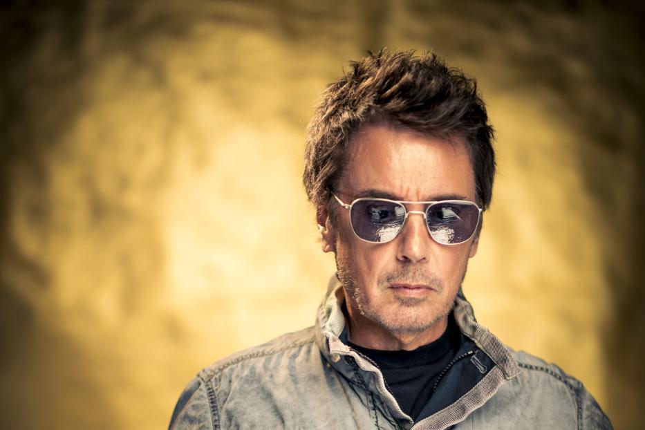 Ein Mann mit Sonnenbrille und grauer Jacke vor einem goldenen Hintergrund.