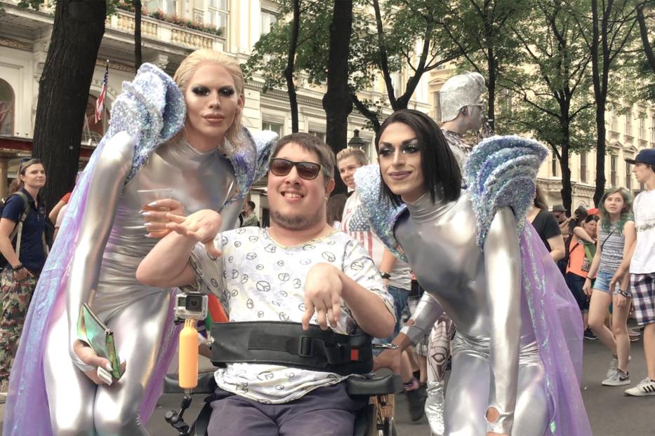 Ein Mann im Rollstuhl posiert mit zwei Dragqueens in silbernen Kostümen für ein Foto.
