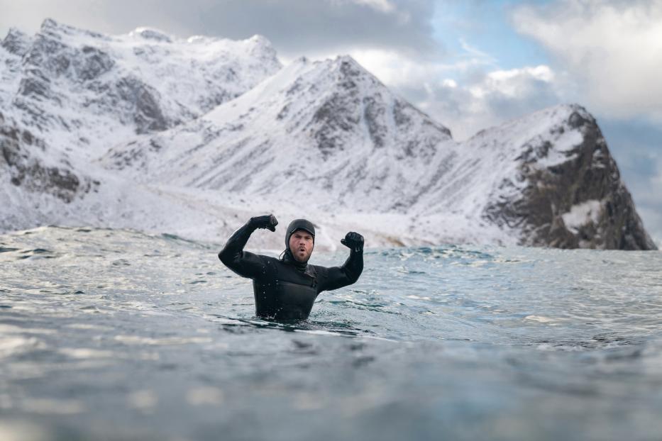 Ein Mann im Neoprenanzug steht im Wasser vor einer schneebedeckten Berglandschaft und ballt die Fäuste.