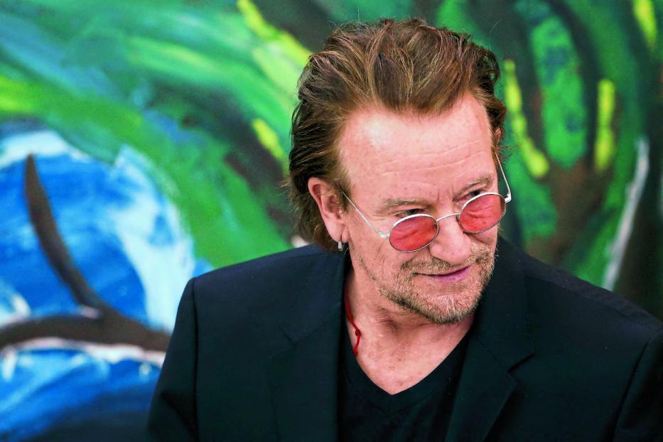 Bono von U2 mit getönter Brille vor einem farbenfrohen Hintergrund.
