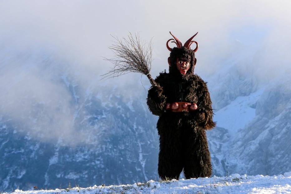 Ein Krampus mit Fellkostüm, Maske und Besen steht vor einer verschneiten Berglandschaft.