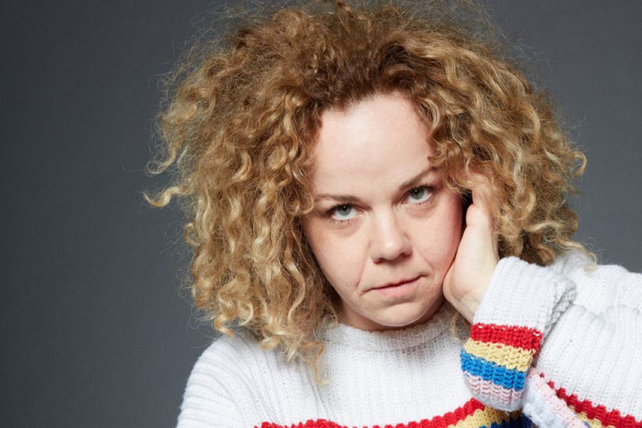 Eine Frau mit lockigem Haar sitzt in Jeans und einem bunten Pullover.