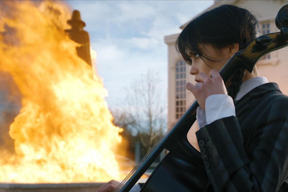 Wednesday Addams spielt Cello vor einem lodernden Feuer.