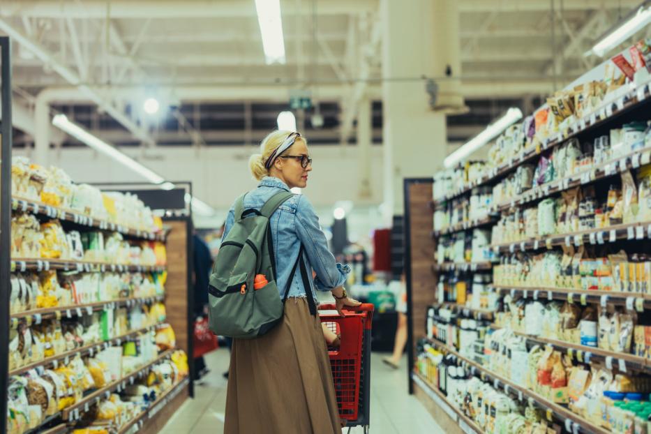 Eine Frau mit Rucksack schiebt einen Einkaufswagen durch einen Supermarkt.