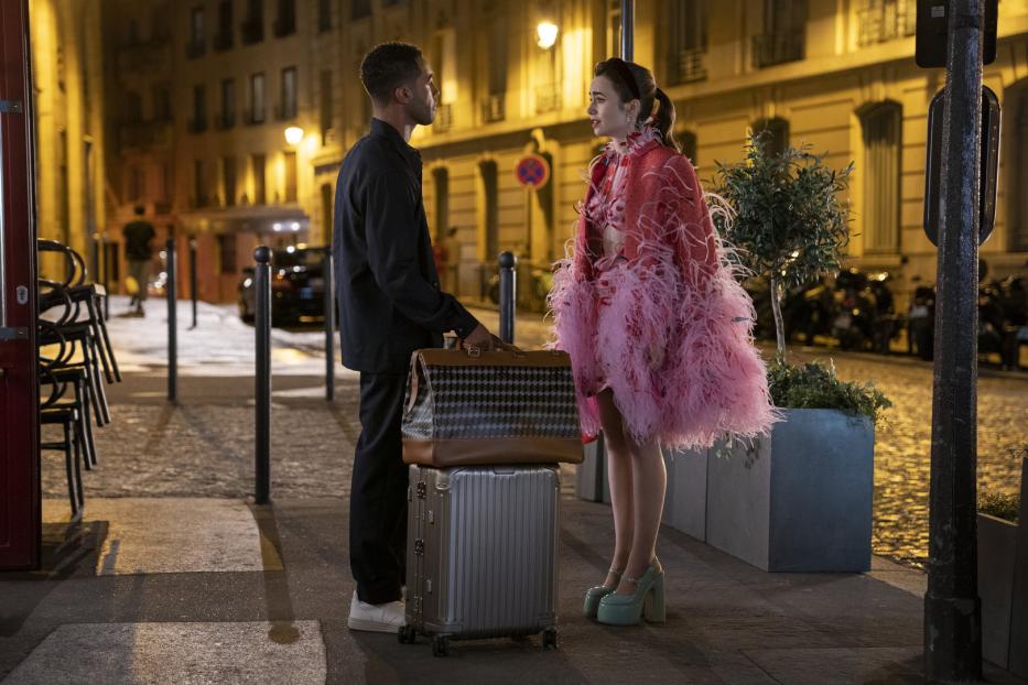 Emily in Paris steht mit Gepäck auf einer Straße und spricht mit einem Mann.