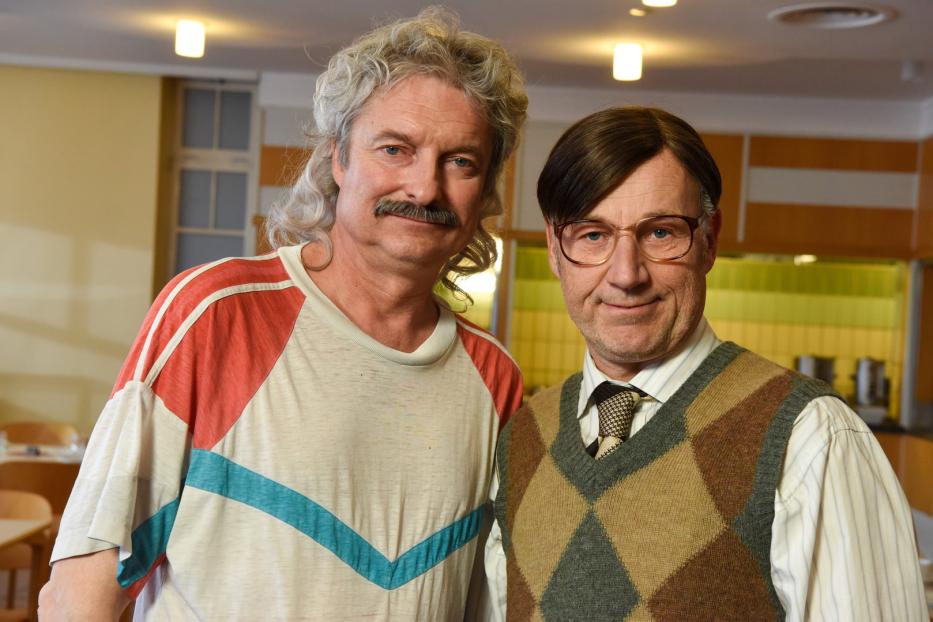 Zwei Männer posieren nebeneinander, der eine mit Schnurrbart und langem, gewelltem Haar, der andere mit Brille und Pullunder.