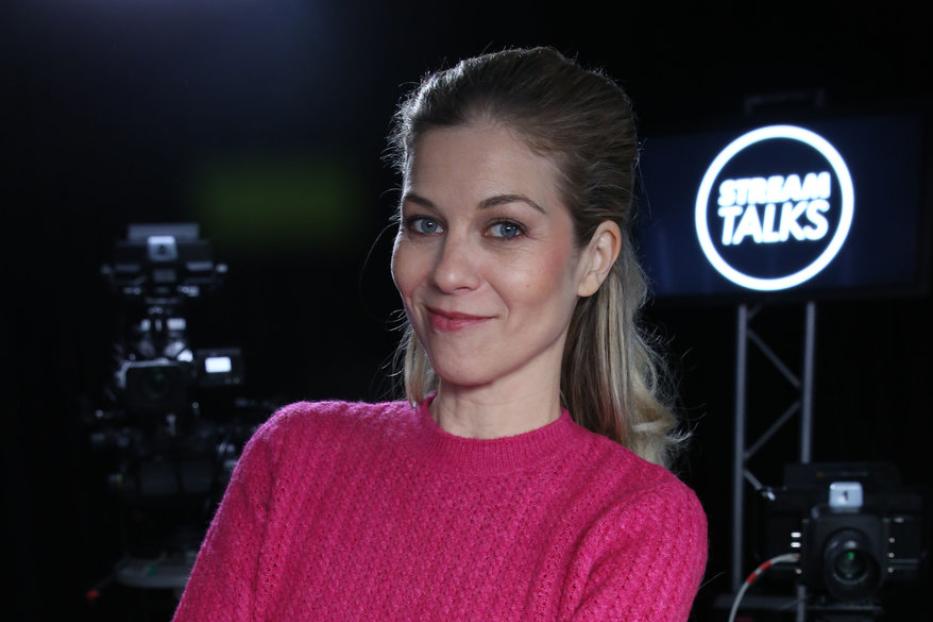 Eine Frau mit verschränkten Armen steht vor Kameras und einem „Stream Talks“-Logo.