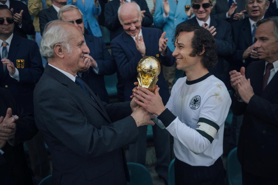 Ein Mann im Anzug überreicht einem Fußballspieler den Weltpokal.