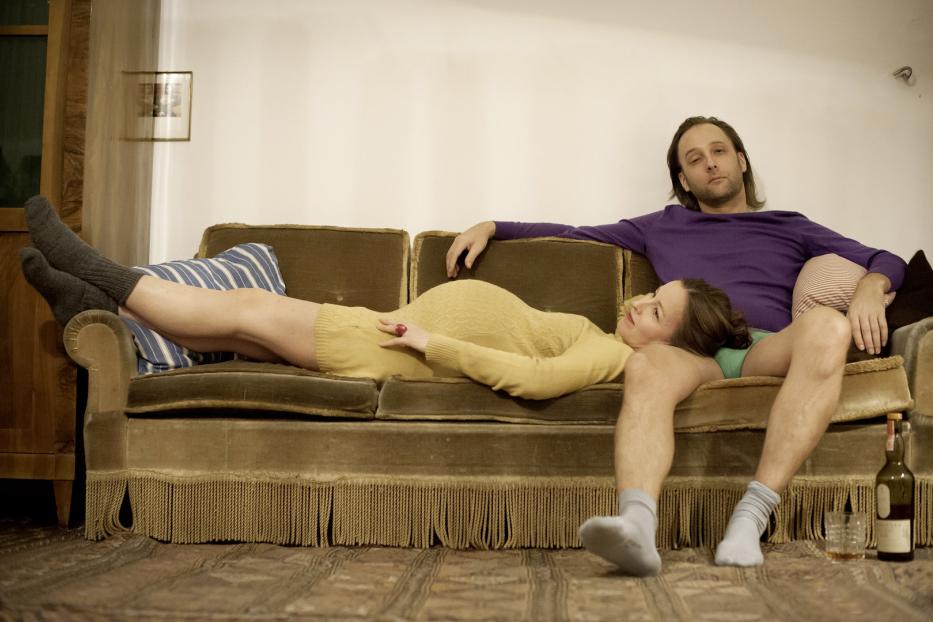 Eine schwangere Frau liegt auf einem Sofa, während ein Mann daneben sitzt.