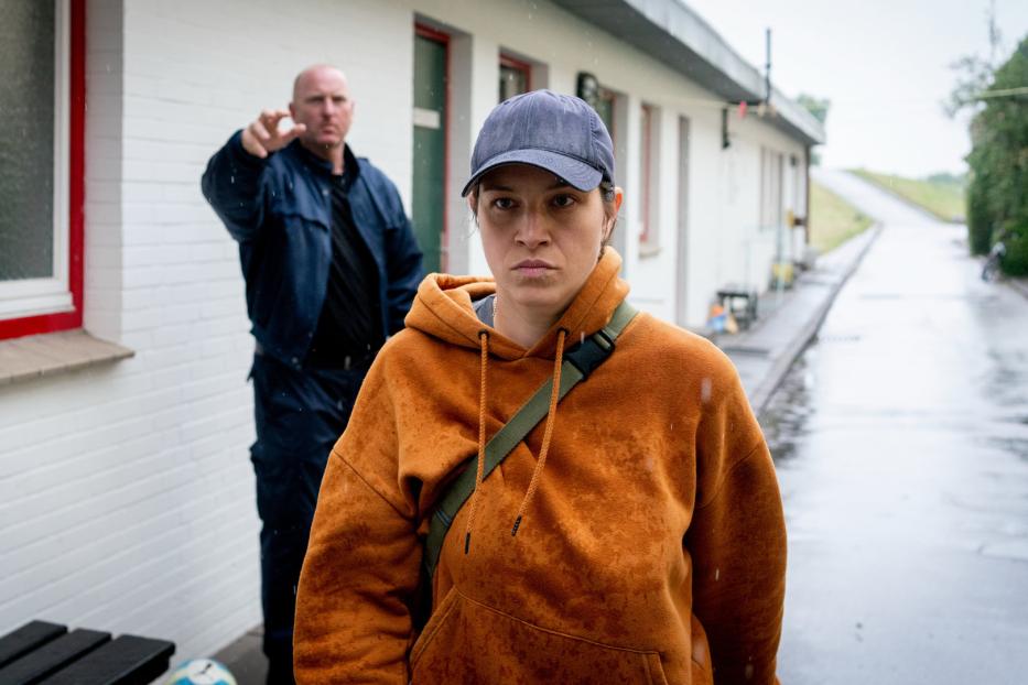 Eine Frau in einem orangefarbenen Hoodie steht im Regen, während ein Mann im Hintergrund auf sie zeigt.