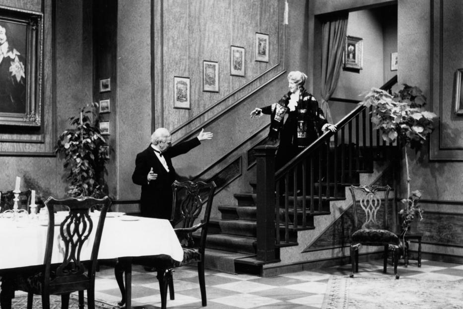 Ein Butler begrüßt eine Dame, die eine Treppe in einem eleganten Haus herunterkommt.