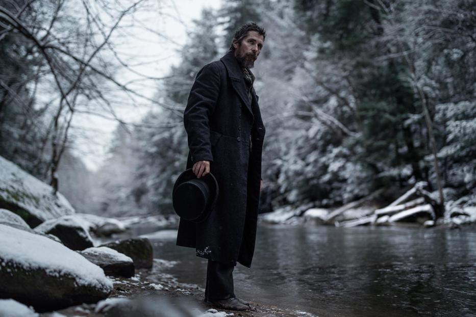 Christian Bale steht in einem langen Mantel an einem Fluss im Winter.