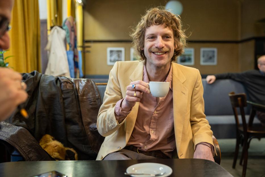 Ein lächelnder Mann mit Schnurrbart sitzt in einem Café und hält eine Tasse in der Hand.