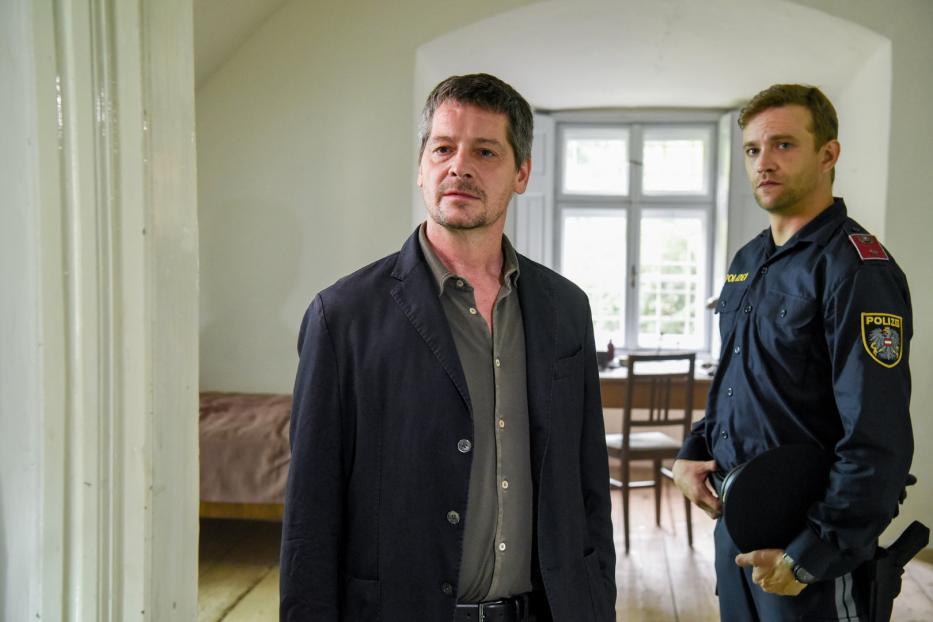 Ein Mann in Zivil steht neben einem österreichischen Polizisten in einem Haus.