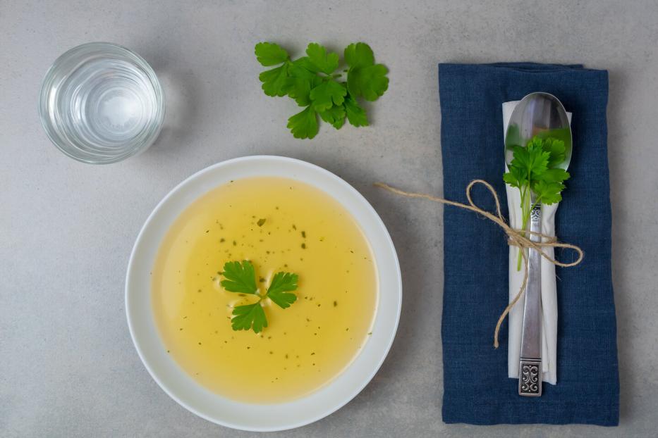 Eine Schüssel mit klarer Brühe, garniert mit Petersilie, neben einem Glas Wasser und einem Besteck auf einer Serviette.