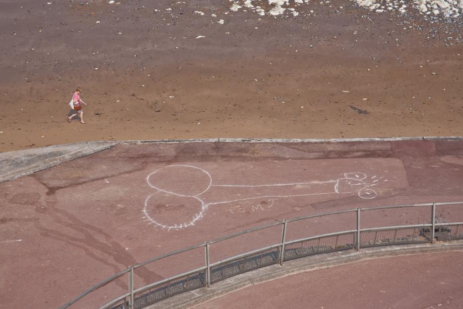 Eine Frau geht am Strand entlang; auf dem Gehweg ist eine Zeichnung mit Kreide zu sehen.