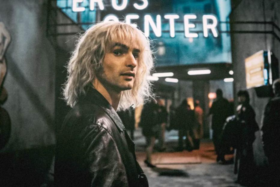 Ein Mann mit blonden Haaren steht vor einem Gebäude mit der Aufschrift „Eros Center“.